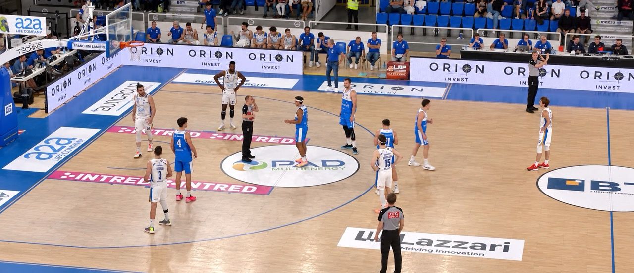 Partita di basket tra squadre in divise bianche e blu sul campo, sponsor Orion visibile.